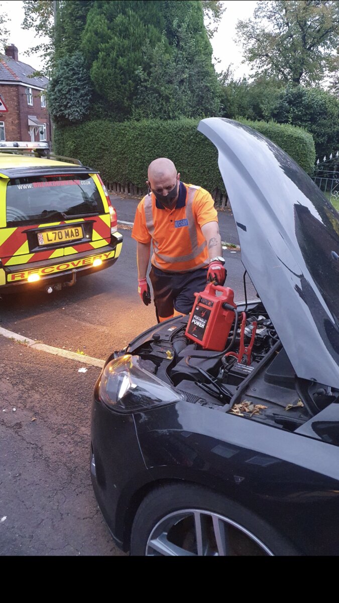 Simon jump-starting a flat battery on a black car with the Breakdown Man recovery van behind on a residential street.