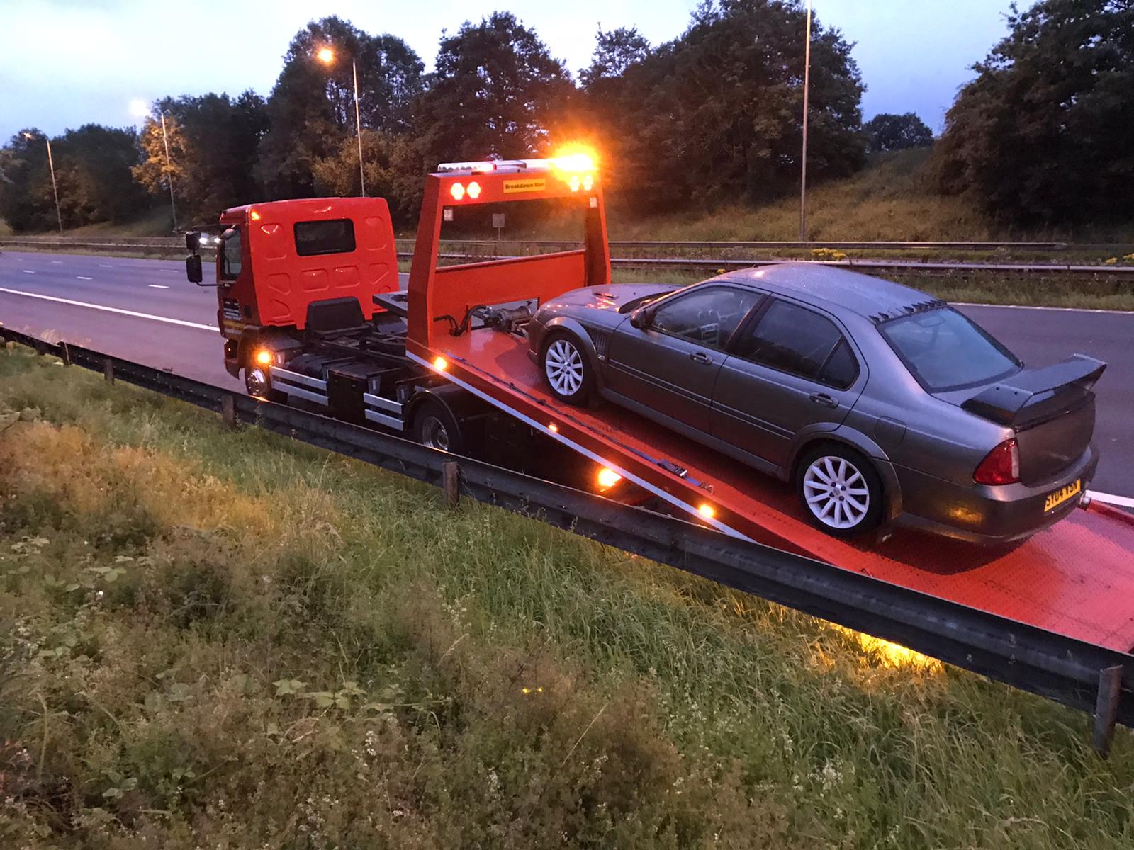 Breakdown Man operator in red high-vis crouched at the front wheel of a grey Audi A6 saloon as it loads onto the Breakdown Man recovery truck on a Wigan terraced street