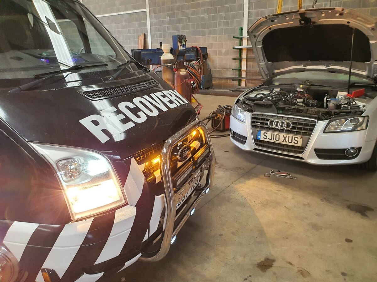 Breakdown Man recovery van beside a silver Audi with bonnet up and jump leads connected for a jump start.