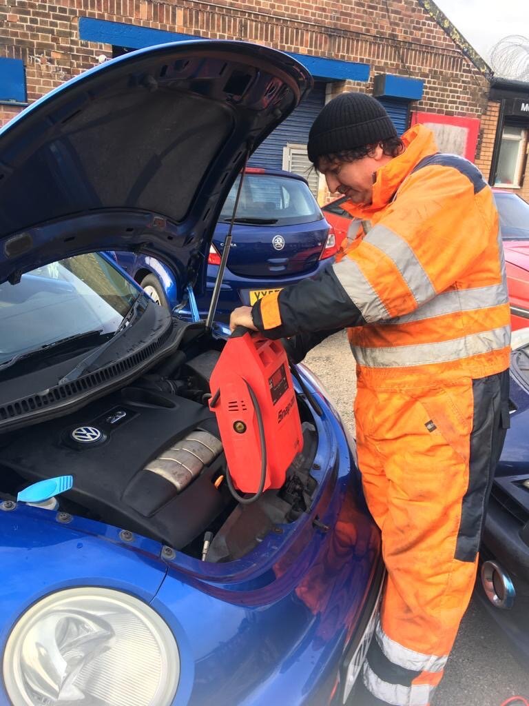 Simon in hi-vis using a red jump pack on a blue VW Beetle during a roadside jump start.
