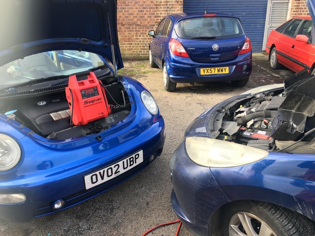 Blue VW Beetle with bonnet up and a red Snap-on jump pack connected during a flat-battery jump start.