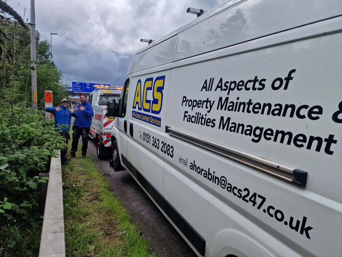 Two recovery operatives stood beside an ACS Property Maintenance van on a grass roadside verge with a Breakdown Man recovery truck behind them.