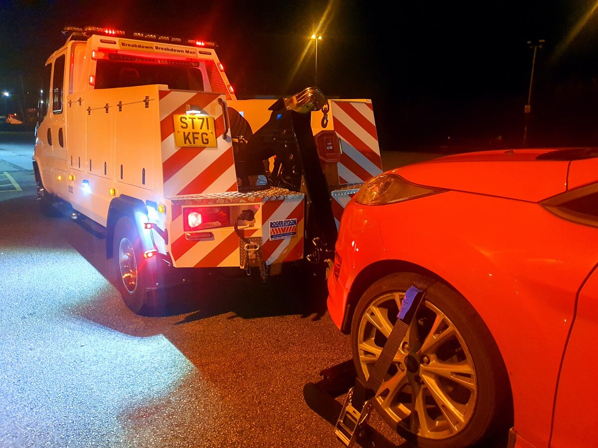 A Breakdown Man recovery truck loading a red car at night while the operator secures it with straps.
