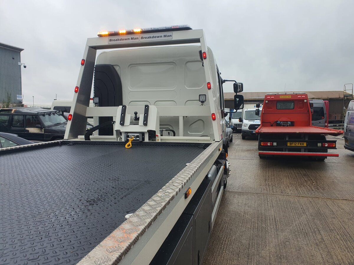 Empty Breakdown Man branded flatbed recovery truck with chequer-plate bed in a yard with a red BF12 FKK recovery lorry parked behind.