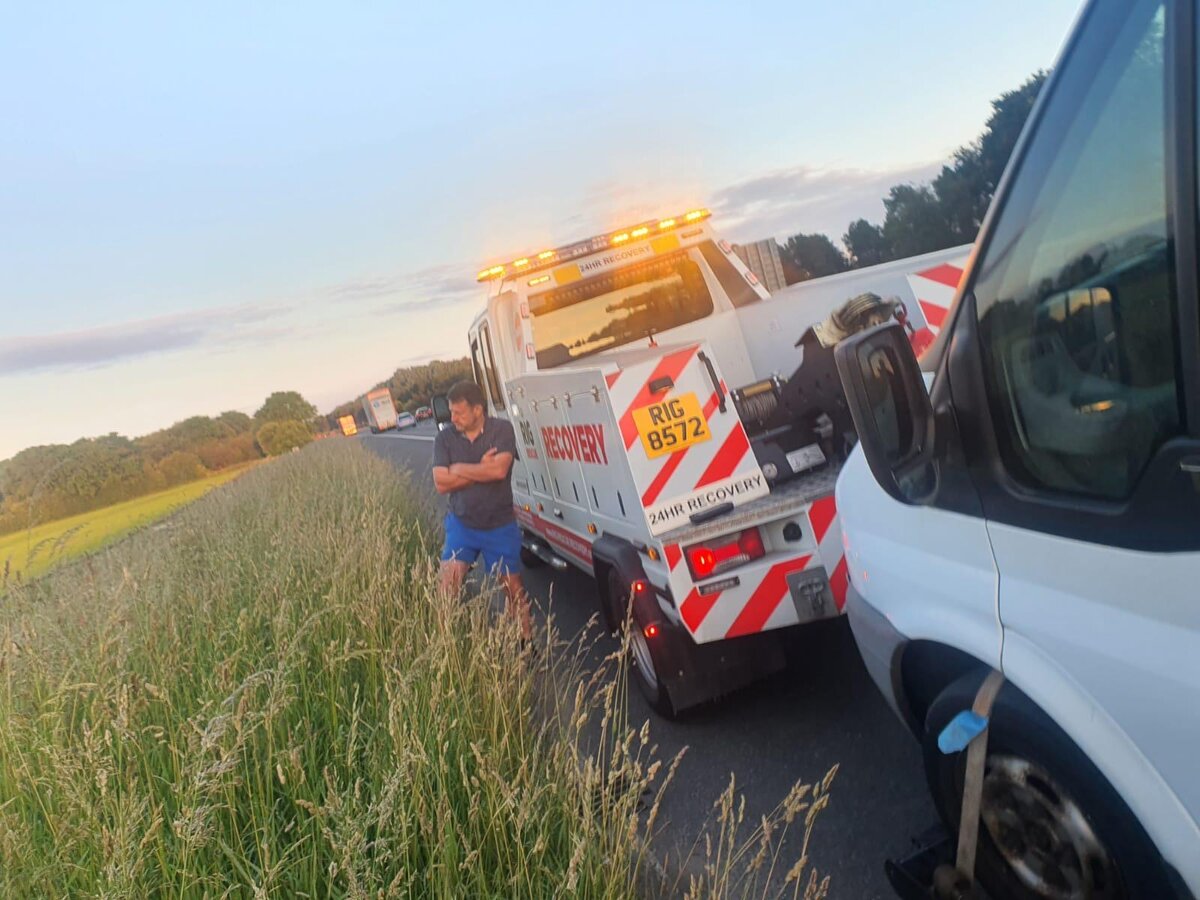 Breakdown Man Rig Recovery 8572 wrecker truck on the A-road verge at sunset, parked in tandem with a white Transit van during a summer-evening recovery.