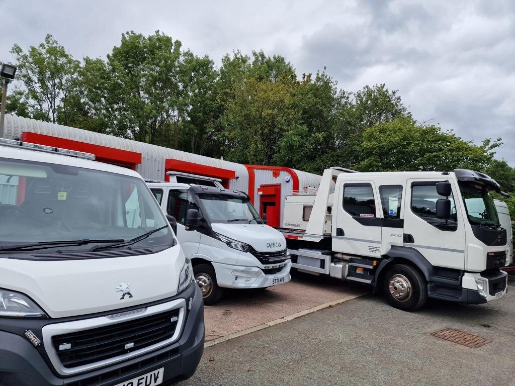 Three Breakdown Man fleet vehicles parked outside the Wigan yard — a white Peugeot Boxer panel van, an Iveco Daily van, and a DAF flatbed recovery truck — under cloudy skies.