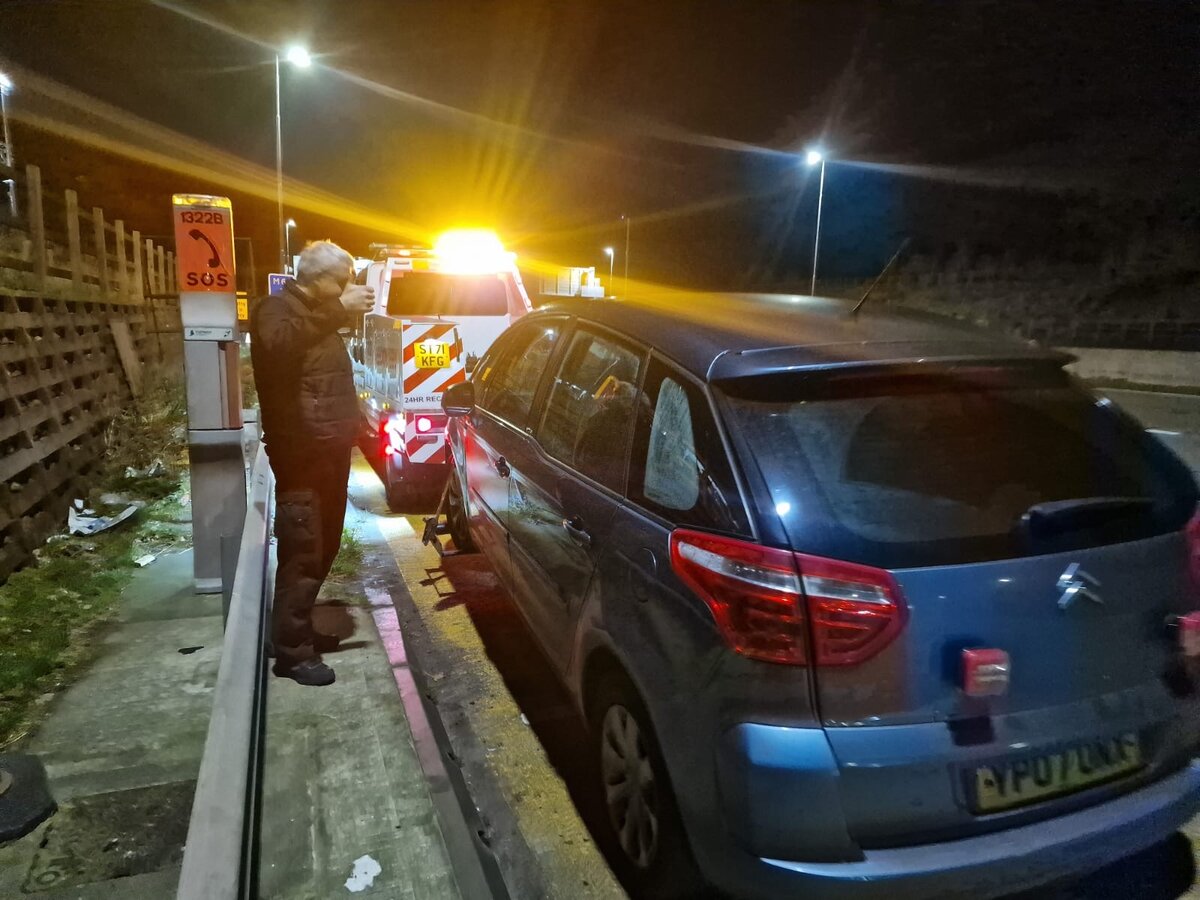 Customer using a motorway SOS emergency phone (post 1322B) at night beside a silver Citroen C4 Picasso, with a Breakdown Man recovery truck flashing amber lights behind.