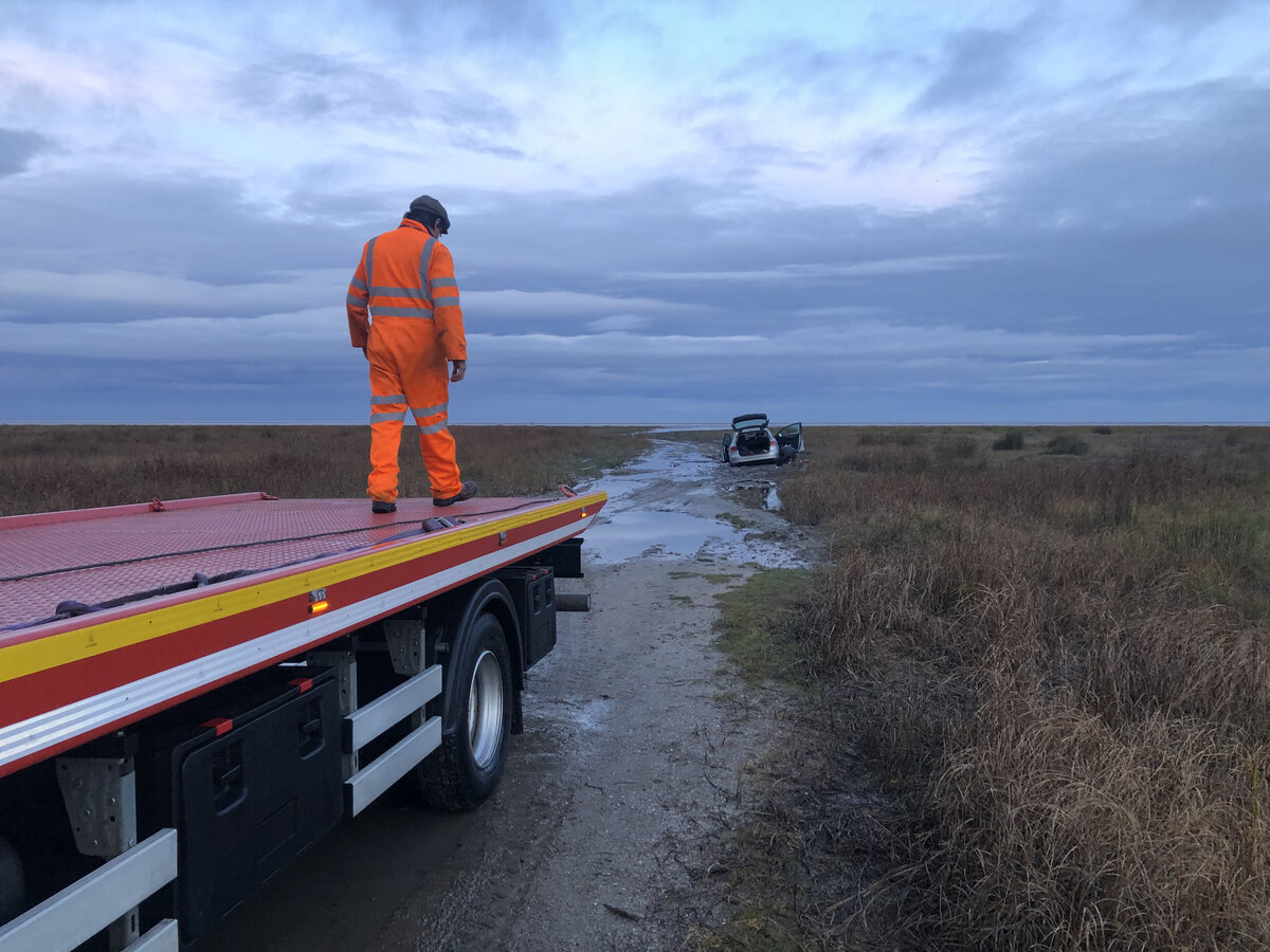 Breakdown Man recovery operator preparing the flatbed at dawn beside a vehicle stranded on a remote marshland track.