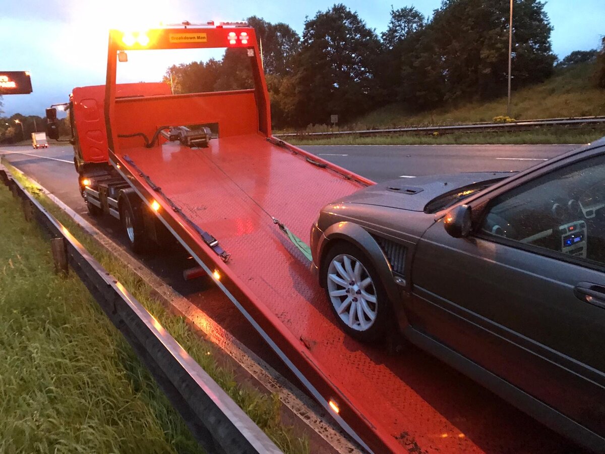 Grey Ford Mondeo loaded onto Breakdown Man's red flatbed at dusk on the motorway with beacon lights running.