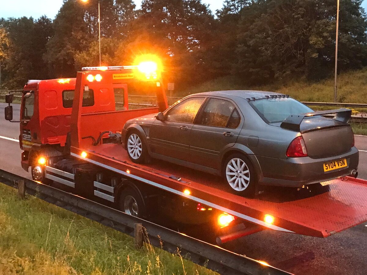 Grey four-door saloon loaded onto a Breakdown Man red flatbed recovery truck at dusk with amber beacons lit beside a motorway carriageway.