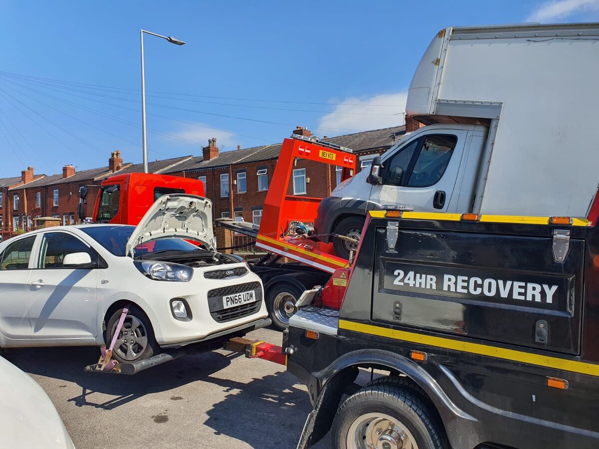 Breakdown Man's 24HR Recovery truck loading a white Kia Picanto with its bonnet up during a roadside recovery on a terraced residential street.