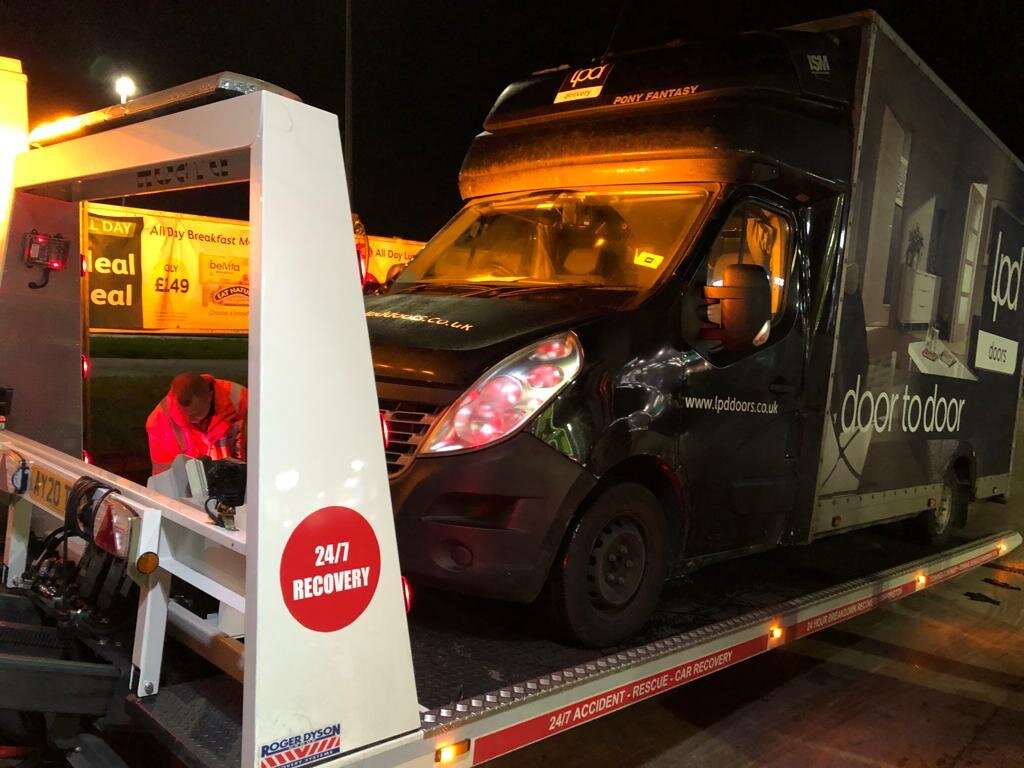Black LPD Doors-branded Renault Master courier van with side-impact damage being loaded onto a 24/7 Recovery flatbed truck at night, recovery operator working alongside.