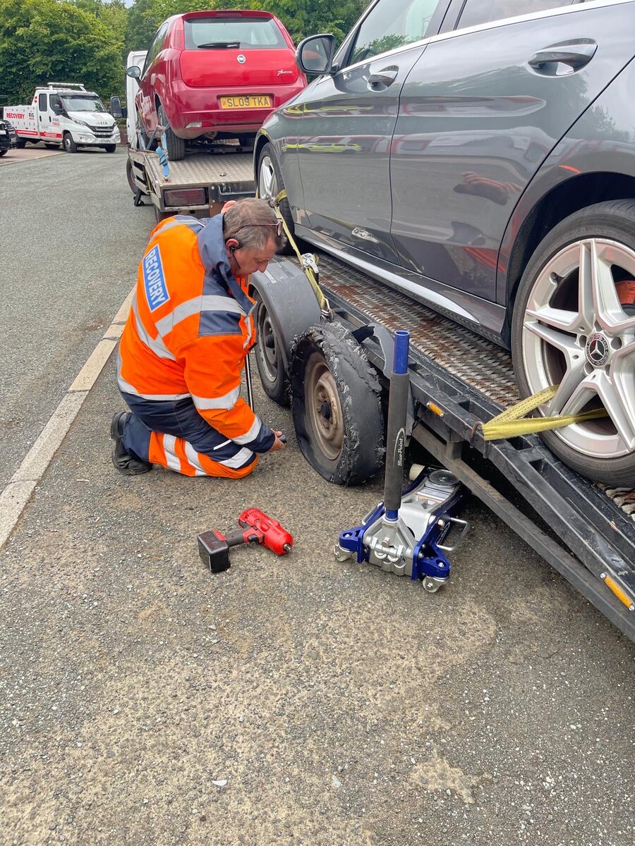 Recovery operator in orange hi-vis strapping a dark Mercedes saloon to the flatbed with a red trolley jack and recovery dollies.