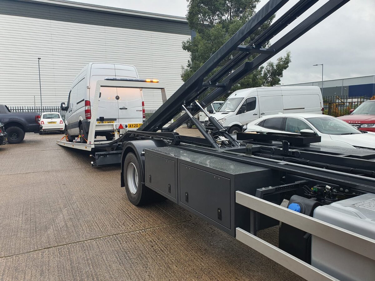 White Mercedes Sprinter panel van loaded onto the Breakdown Man flatbed at an industrial estate, with a second Breakdown Man hook-lift recovery rig in the foreground