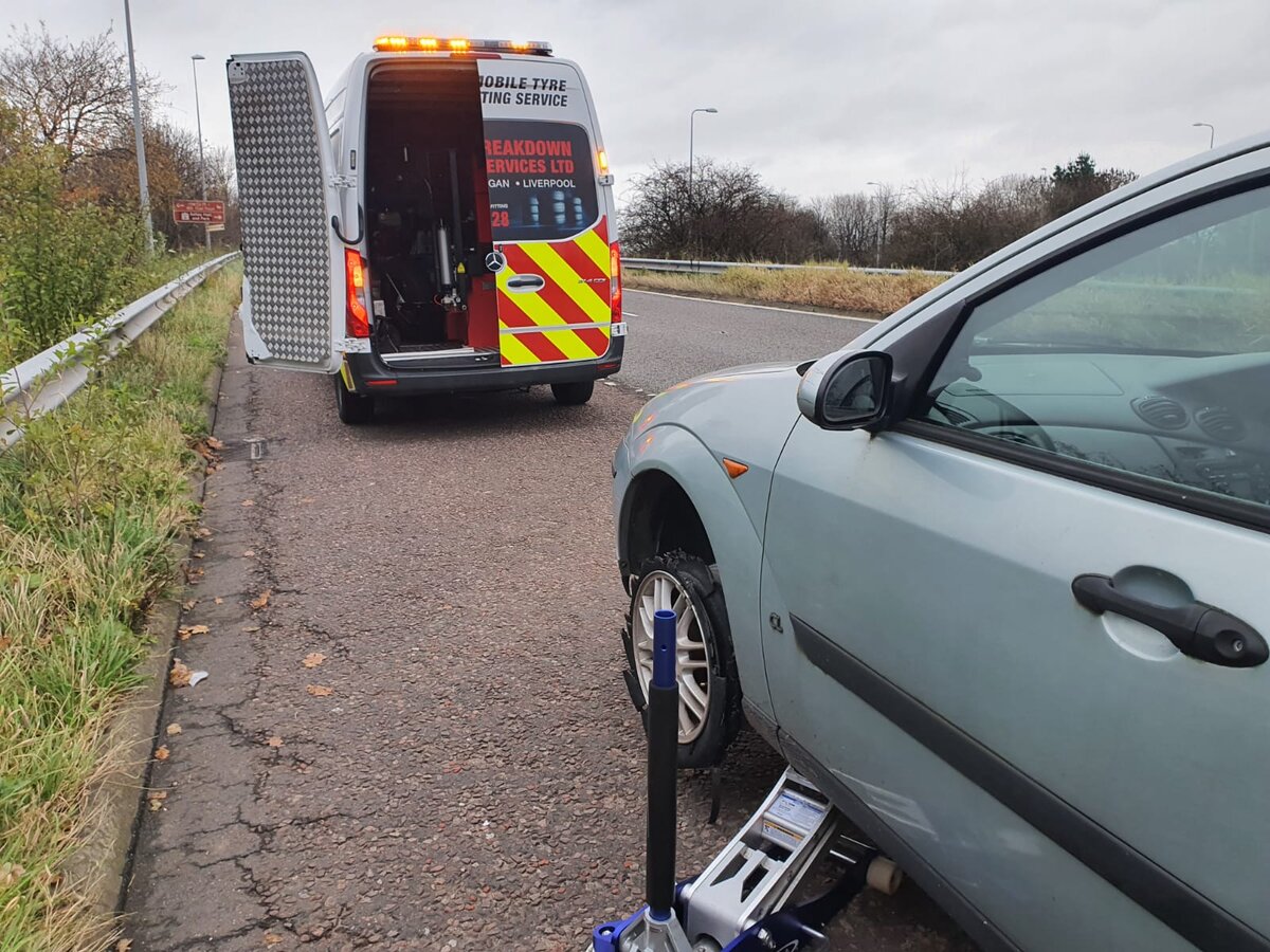 Breakdown Man mobile tyre fitting van with rear ramp deployed, helping a silver Ford Focus with a flat tyre on the roadside.