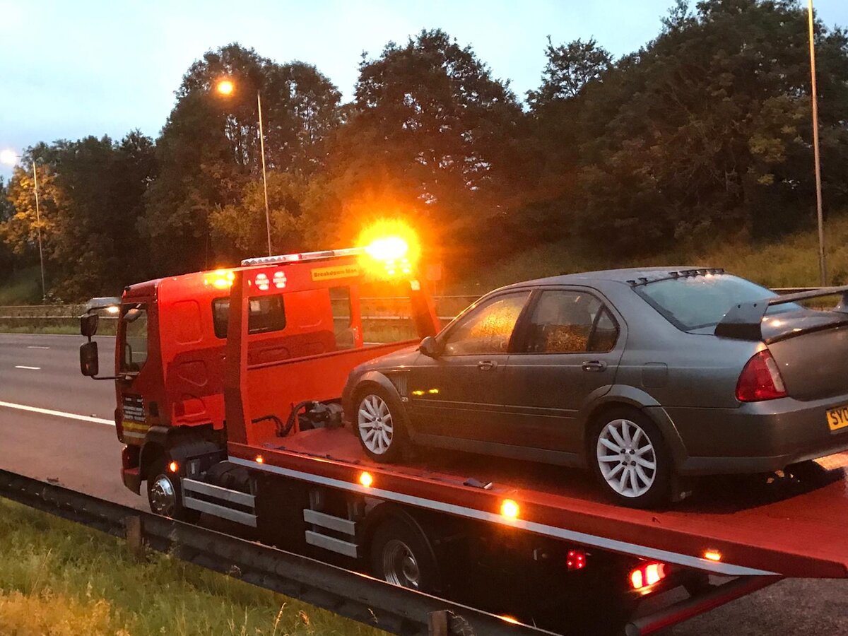 A grey saloon loaded on a Breakdown Man flatbed recovery truck at dusk beside a motorway crash barrier, amber beacon lit.