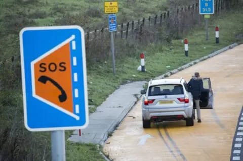 Driver beside a silver SUV in a motorway emergency refuge area marked by an SOS phone sign — Breakdown Man motorway recovery.