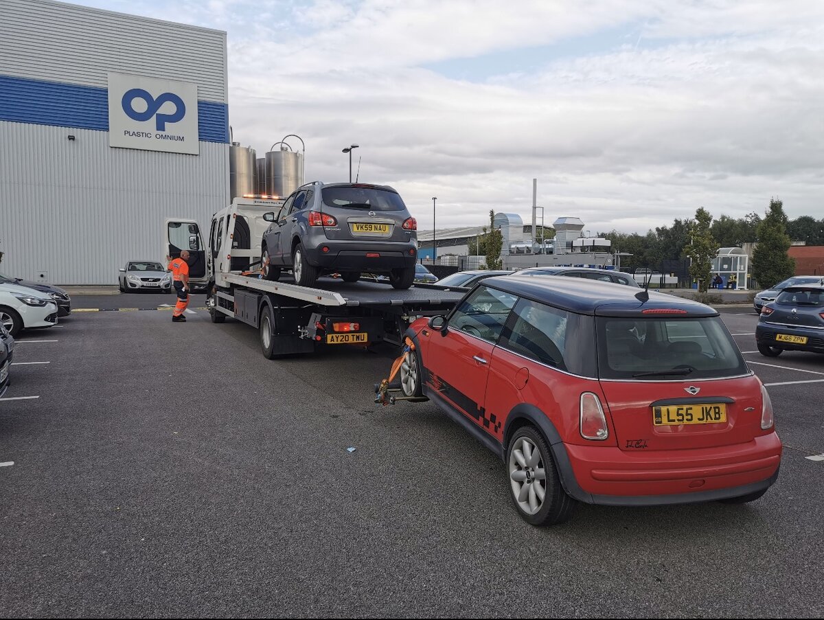 Breakdown Man recovering a grey Nissan Qashqai on the flatbed with a red Mini towed behind, on an industrial site.