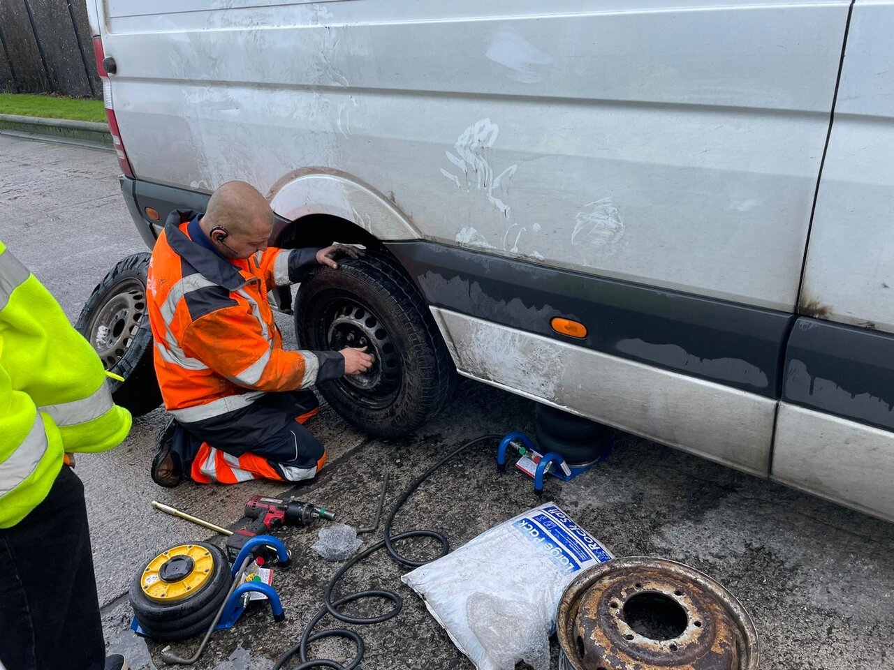 Mobile tyre fitter in orange hi-vis kneeling at the rear wheel of a white Sprinter van, fitting a replacement tyre on the roadside