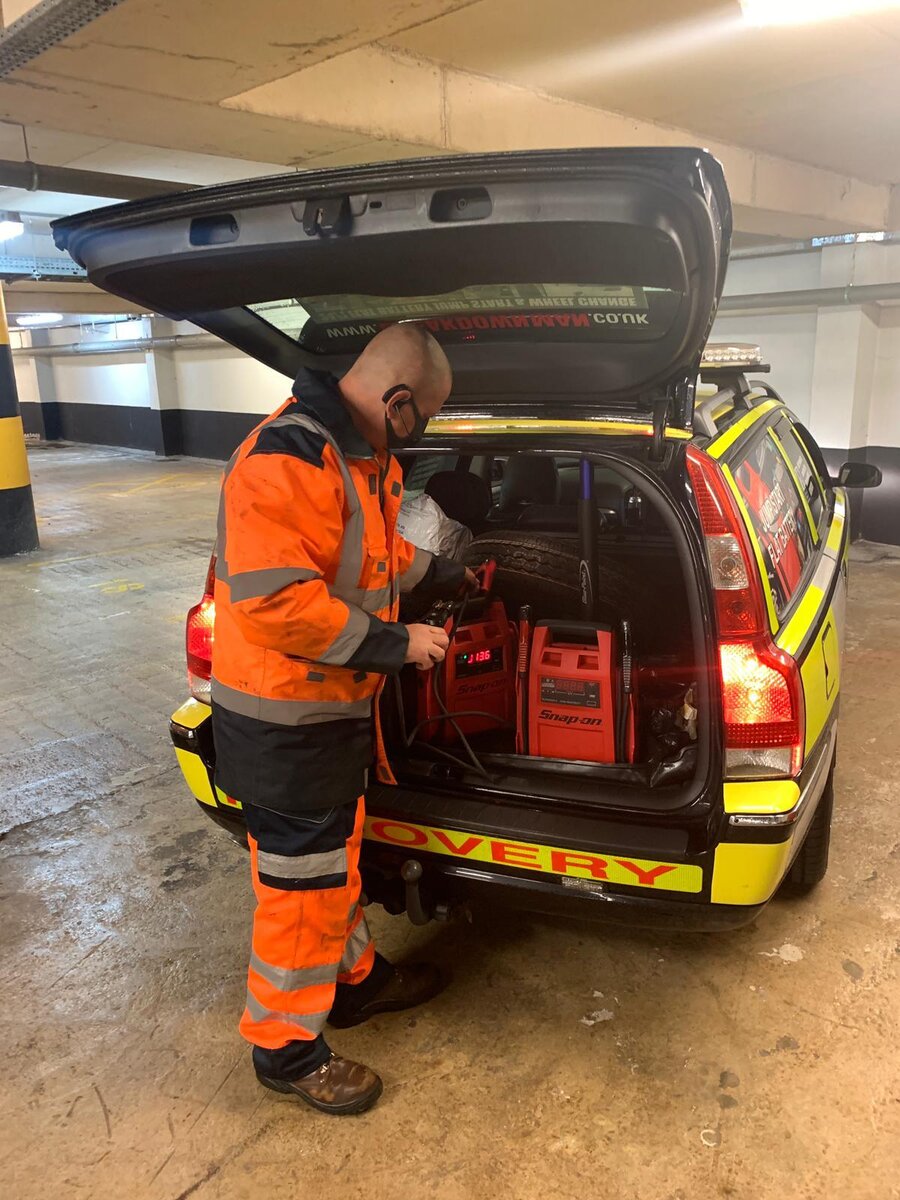 Breakdown Man operator in orange hi-vis using a Snap-On Booster Pac to jump-start a stranded Volvo V70 highway-maintenance vehicle in a Skelmersdale car park.