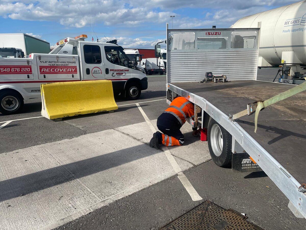 Breakdown Man operator in orange hi-vis kneeling under a flatbed trailer at the Wigan depot to fit straps before a St Helens job.