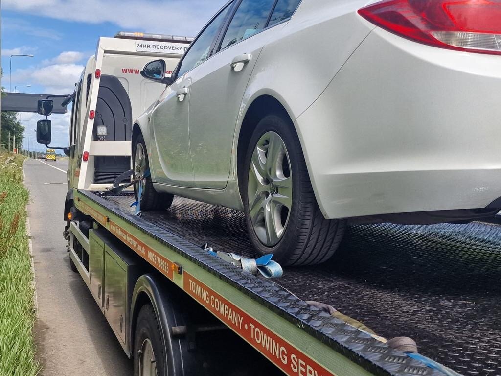 White Vauxhall Insignia saloon strapped down on a Breakdown Man 24HR Recovery flatbed truck on a rural roadside verge.