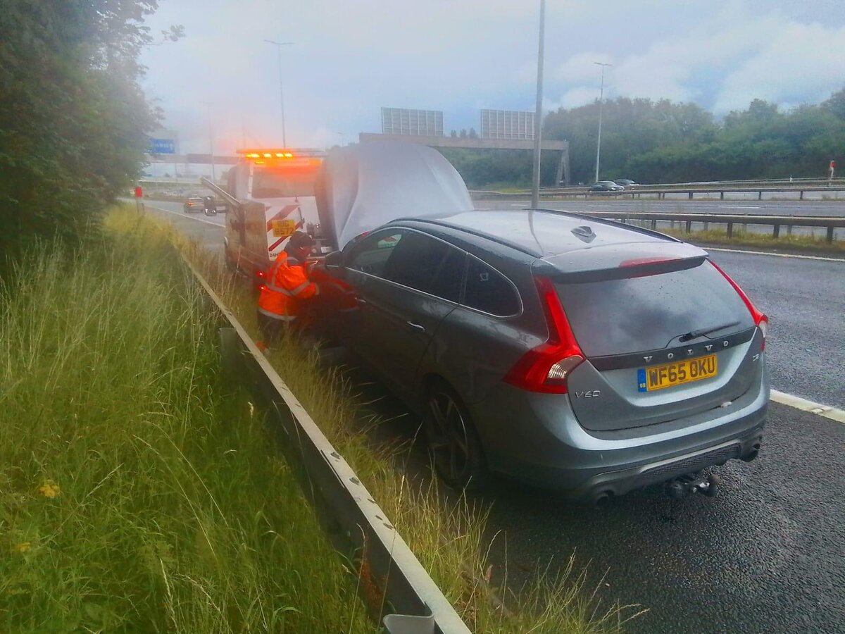 Silver Volvo V60 estate with bonnet up on the motorway hard shoulder, Breakdown Man's recovery vehicle attending behind the barrier.