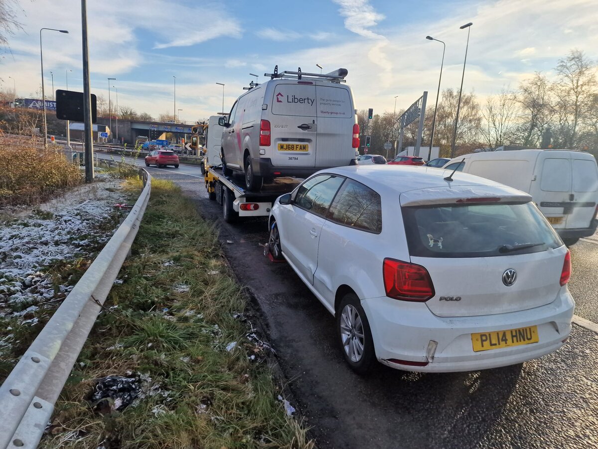 White Volkswagen Polo parked on a frost-dusted roadside verge alongside a Liberty-branded service van and a Breakdown Man flatbed recovery truck on the slip road.