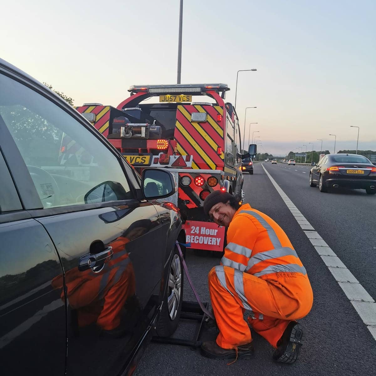 Breakdown Man operator in hi-vis crouching to strap a car onto the recovery truck on a motorway hard shoulder at dusk, with traffic passing.