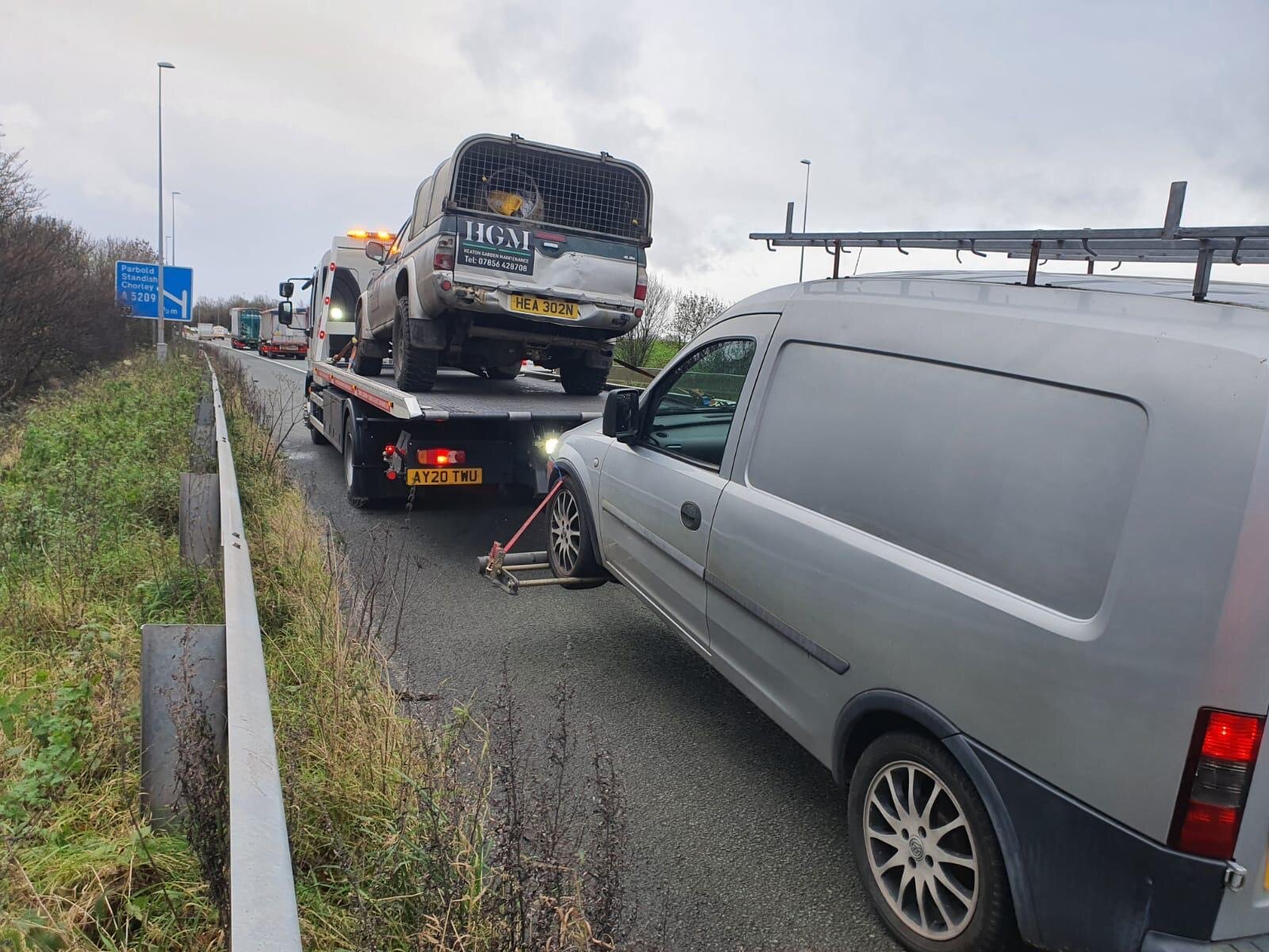 A Land Rover Defender loaded onto a flatbed recovery truck on the M6 hard shoulder, alongside a stranded grey van waiting to be cleared.